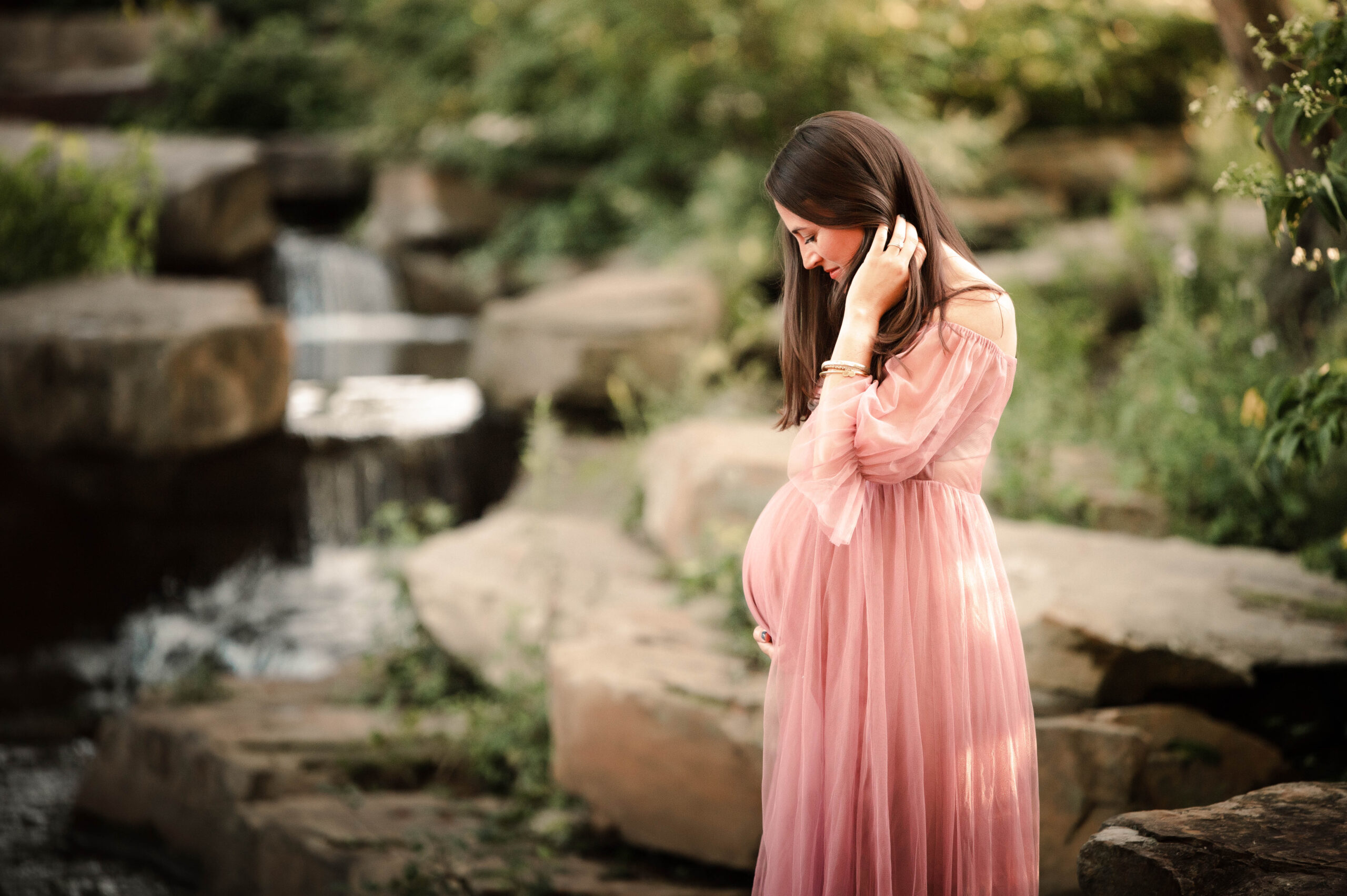 Maternity photo of pregnant woman standing next to creek in a pink gown utilizing services of a doula in dayton, oh