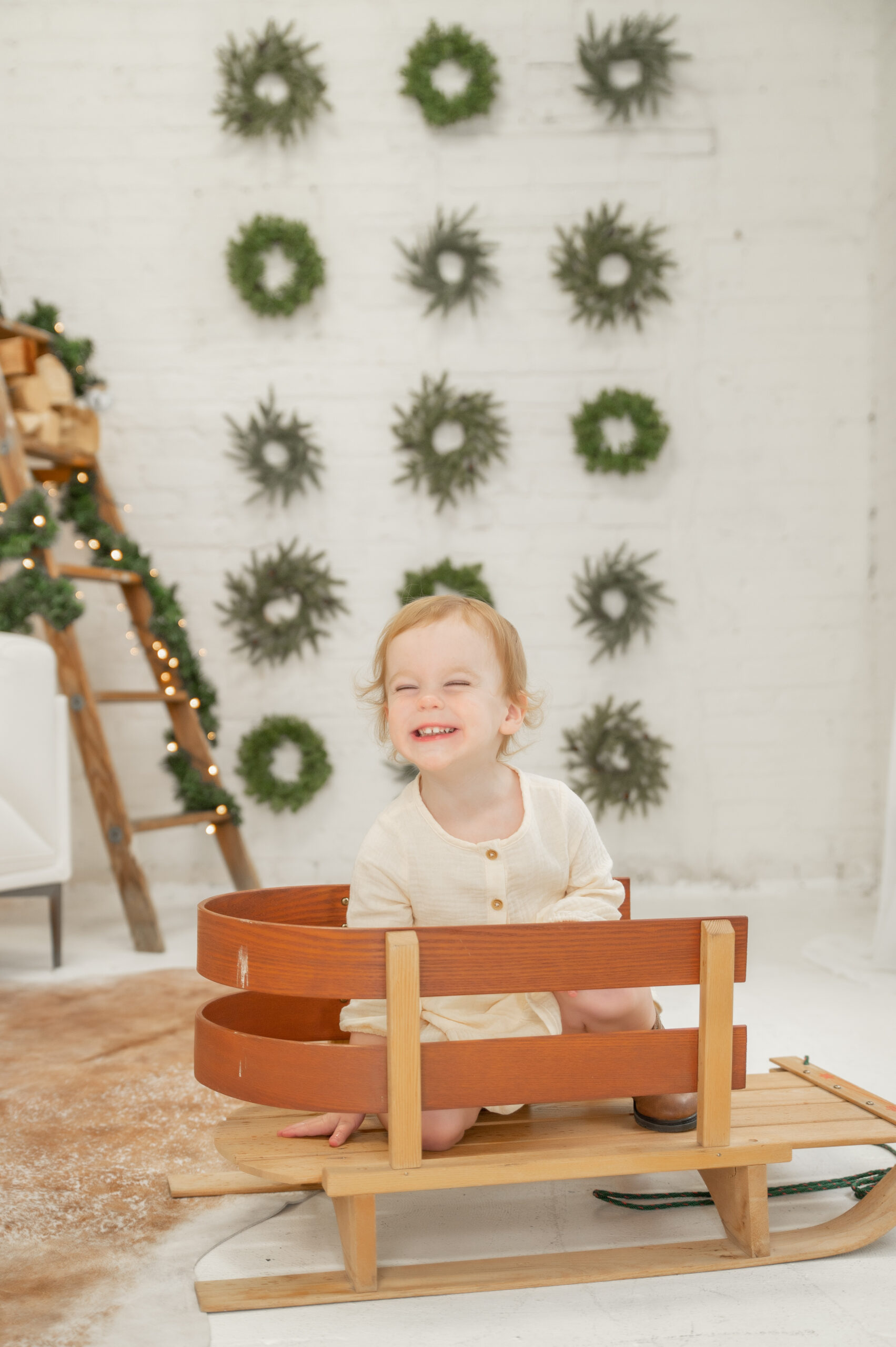 Toddler sitting in Christmas sleigh with wreath backdrop and a big smile on her face!