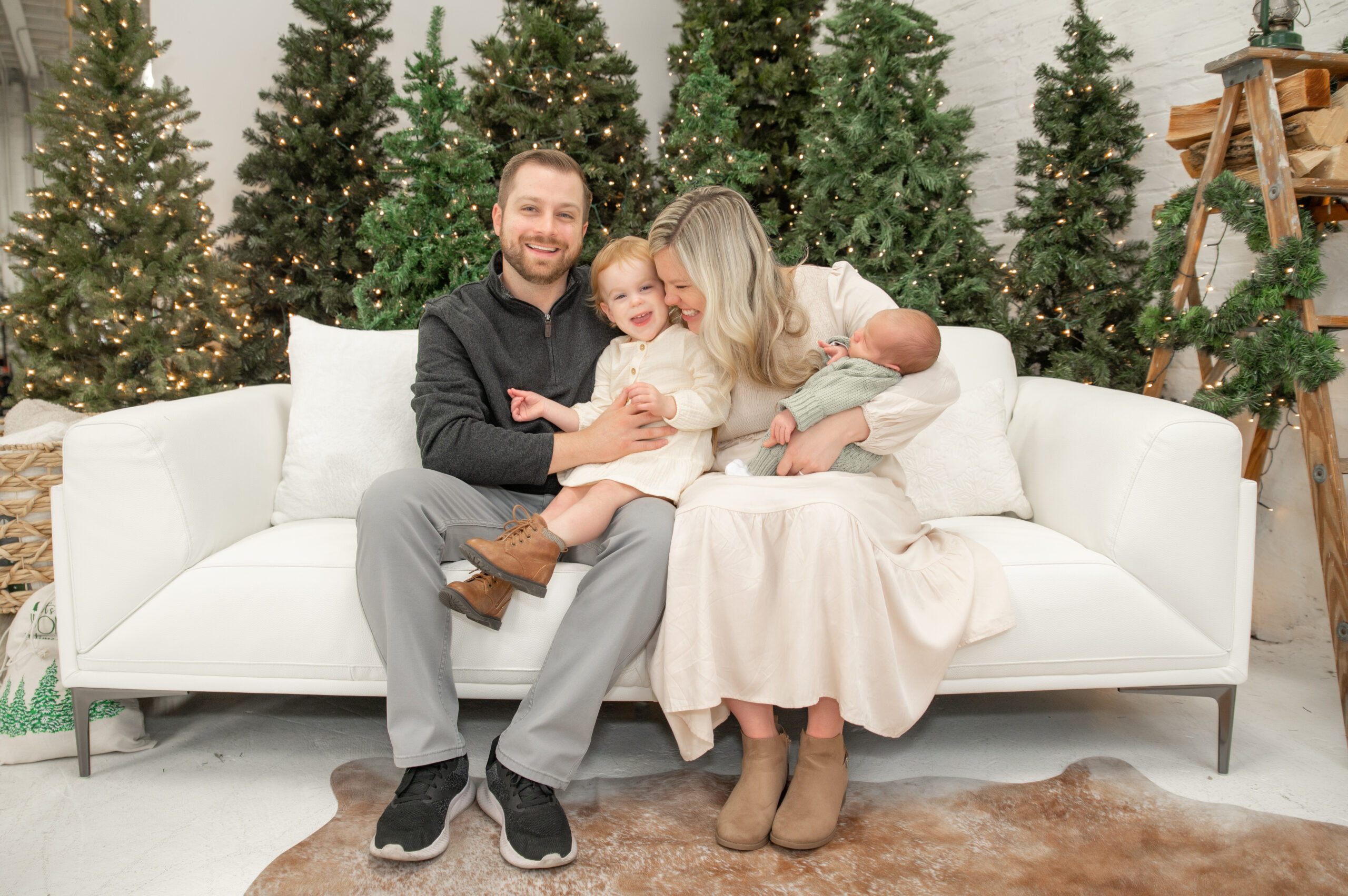 Family cuddled on couch in front of Christmas trees with newborn.